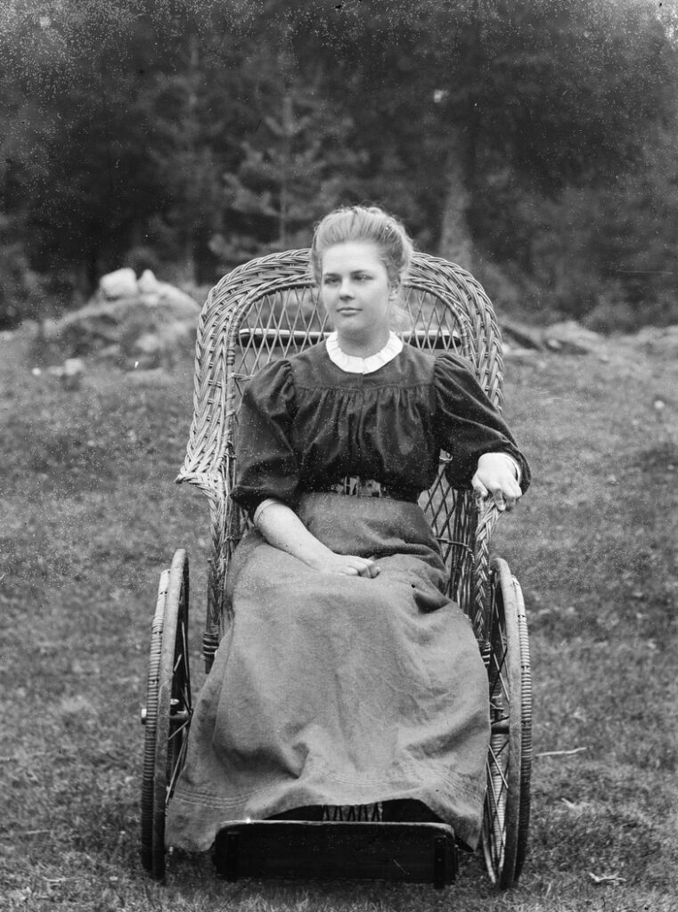 Ung kvinna sittandes i korgrullstol år 1910. Hon är klädd i blus och lång kjol. Håret är uppsatt.