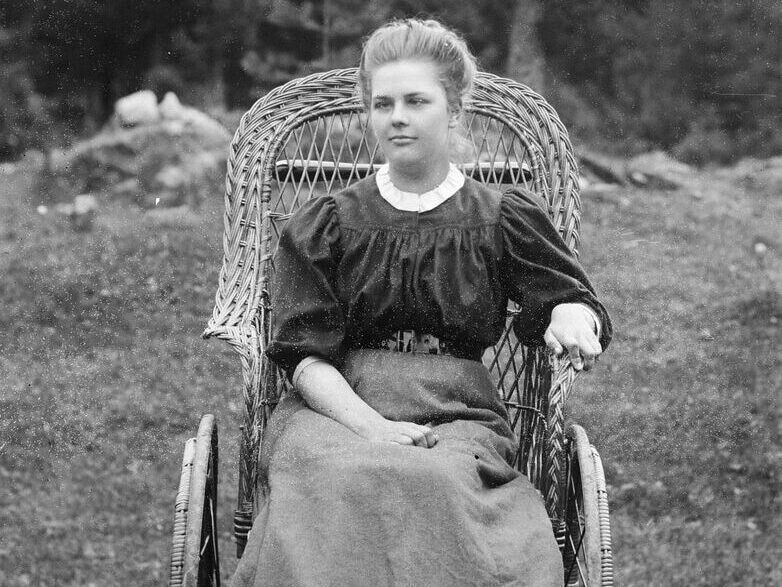 Ung kvinna sittandes i korgrullstol år 1910. Hon är klädd i blus och lång kjol. Håret är uppsatt.